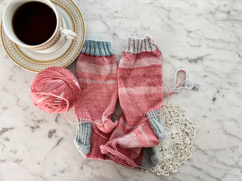 Two pink socks with blue cuffs, heels, and toes are laid out on a white marble countertop. The right sock is still on the needles and needs a finished toe. There is a gold and white cup full of tea and a vintage doily next to the socks.
