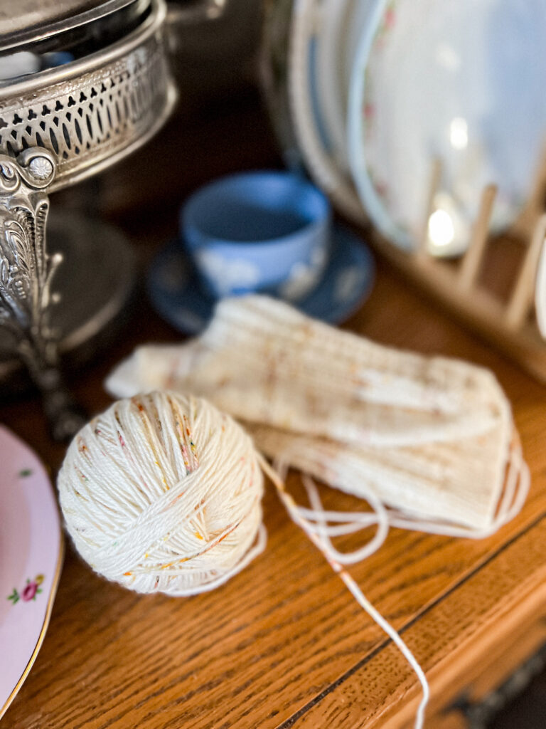 A ball of cream-colored yarn with soft speckles sits on an old oak china hutch surrounded by vintage dishes and a finished sock that needs its ends woven in.