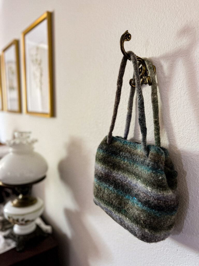 A striped, knit bag in shades of green, gray, and purple hangs from an ornate brass hook. The bag is knit and then felted.