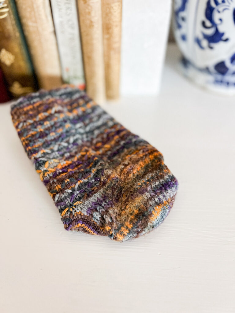 A purple, blue, and orange knit sock is folded on a white dresser top so that the felted heel is visible.