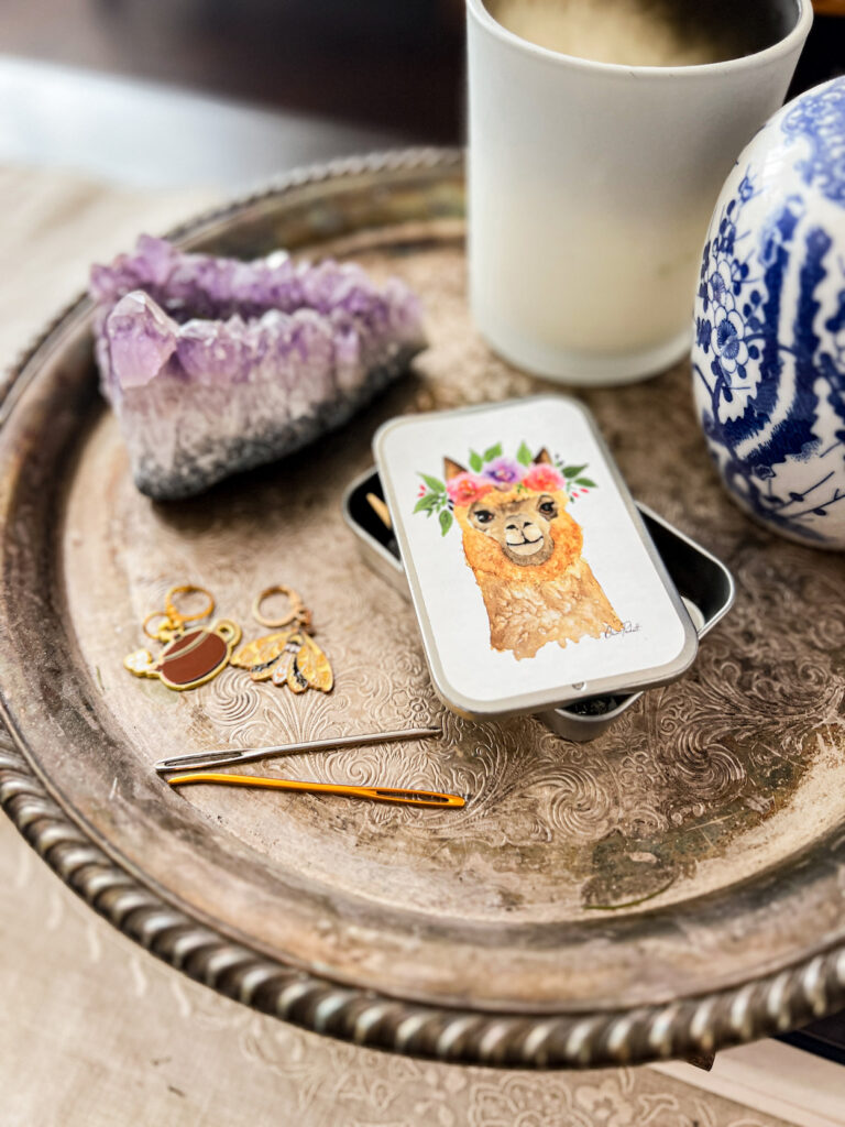 Some enamel stitch markers (one a brown teapot with steam coming from its spout, one a yellow and black moth) sit on a silver tray next to a couple darning needles and a notions tin with a sliding lid (there's an illustration of an alpaca on the lid).