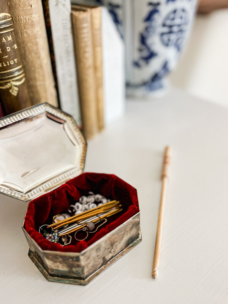 A silver box with red velvet lining holds lots of stitch markers, darning needles, and progress keepers. On the white tabletop next to it is a wooden crochet hook.