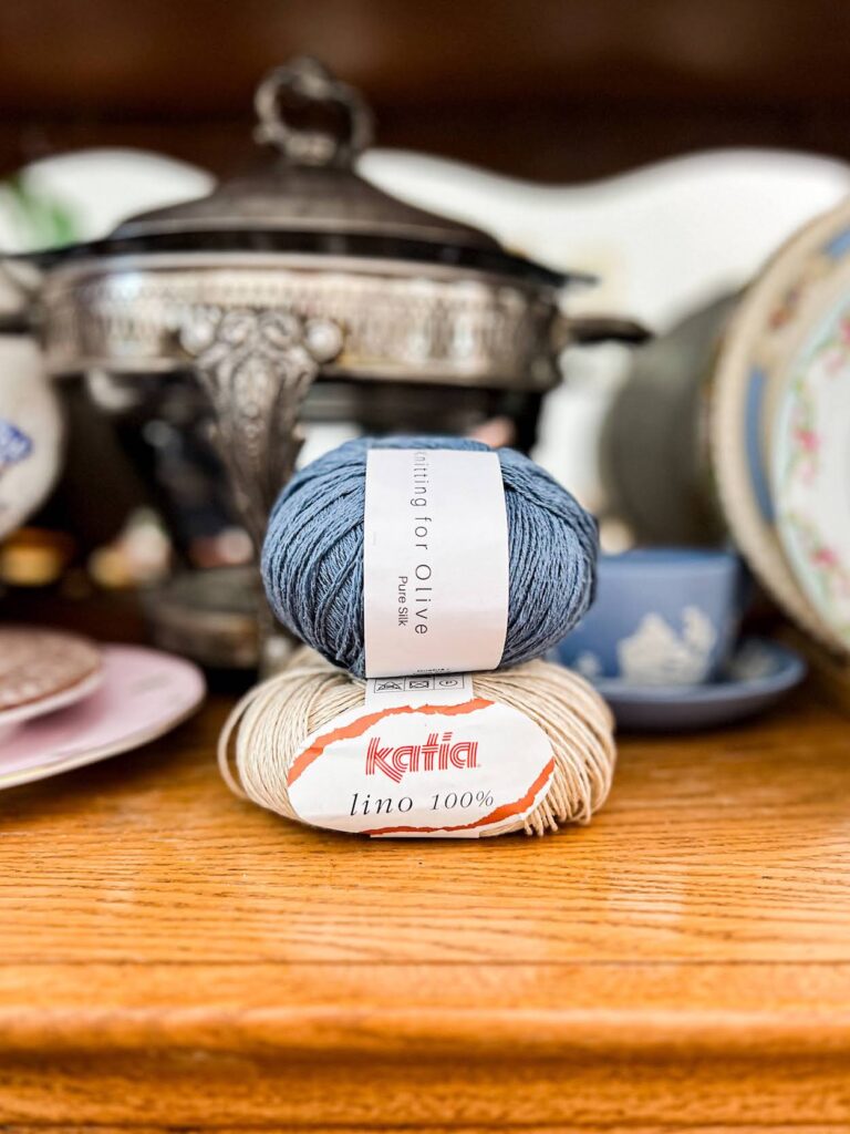 A blue ball of silk yarn is stacked on top of a tan ball of linen yarn and photographed from the front, surrounded by lots of antique dishes on the shelf of an old oak china hutch.