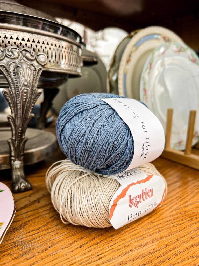 A blue ball of silk yarn is stacked on top of a tan ball of linen yarn and photographed from the side, surrounded by lots of antique dishes on the shelf of an old oak china hutch.