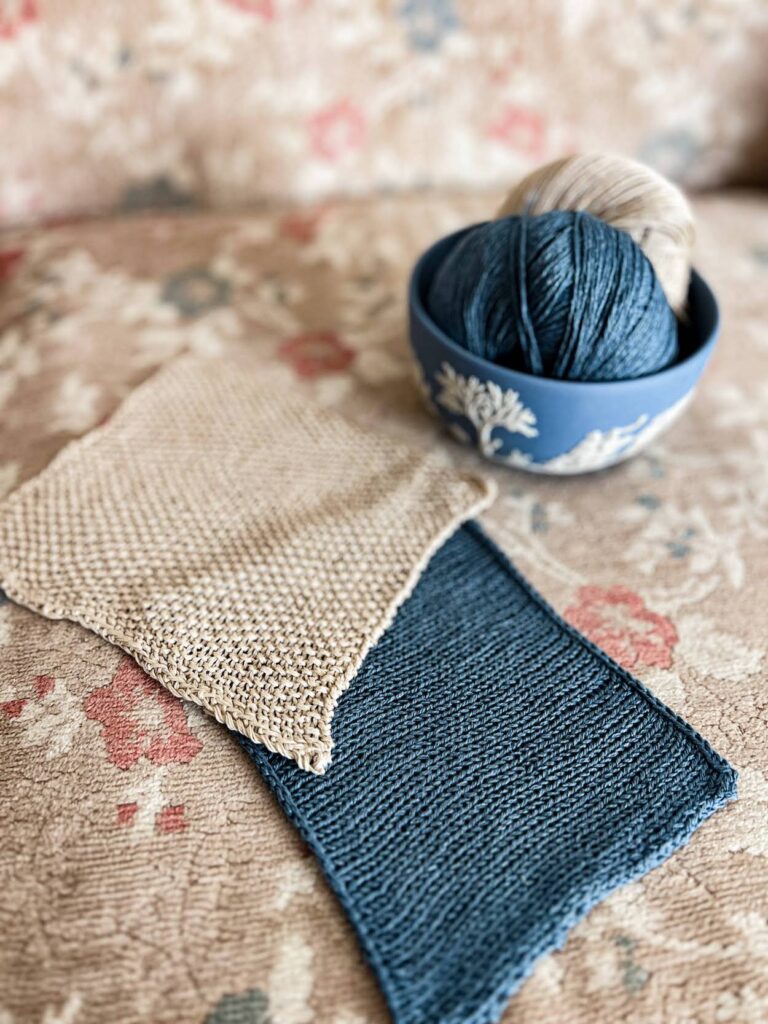 Two knit swatches, one in tan linen seed stitch and one in blue silk stockinette, rest on a vintage sofa with faded floral upholstery. At the top right is a blue and white Jasperware bowl with two balls of yarn in it that match the swatches.
