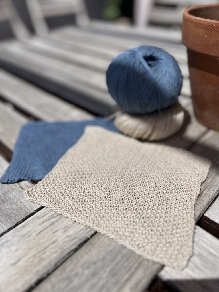 A low-angle photo of two knit swatched, one in blue silk stockinette and one in tan linen seed stitch, on a worn teak table in bright sunshine. Blurred in the background is a small stack of balls of yarn in the same colors and materials as the swatches.
