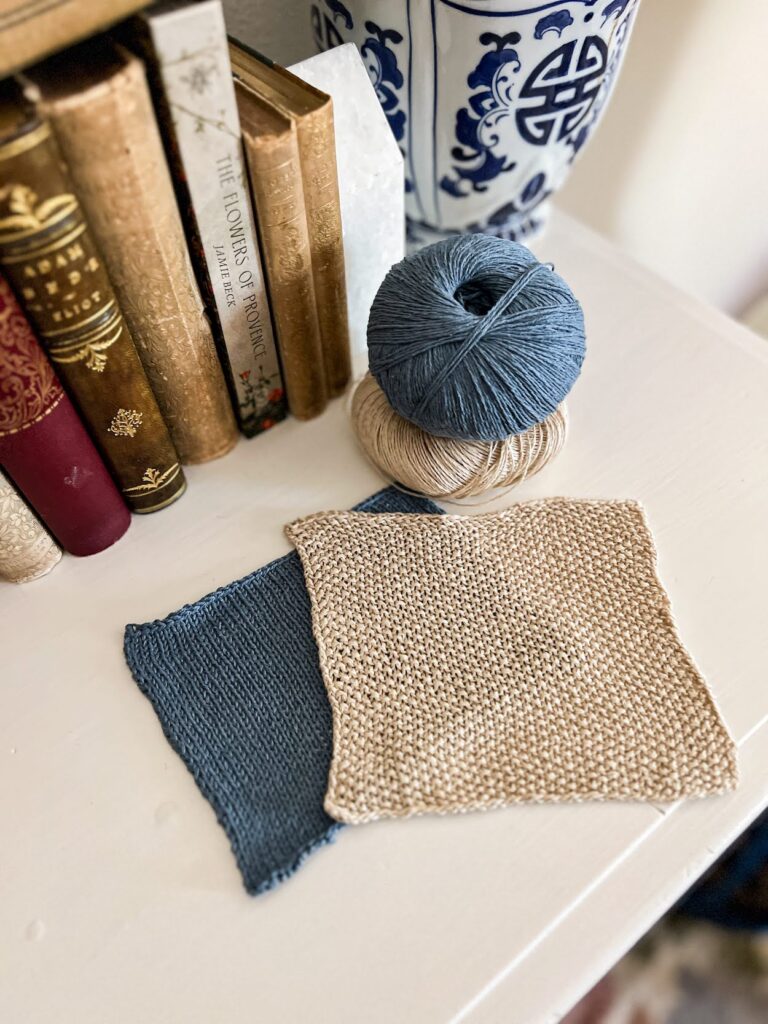 A pair of knit swatches, one in blue silk stockinette and one in tan linen seed stitch, rest on top of a white dresser next to a small stack of yarn balls in the same colors and materials. To the top left are several antique books and a blue and white pot for an orchid.