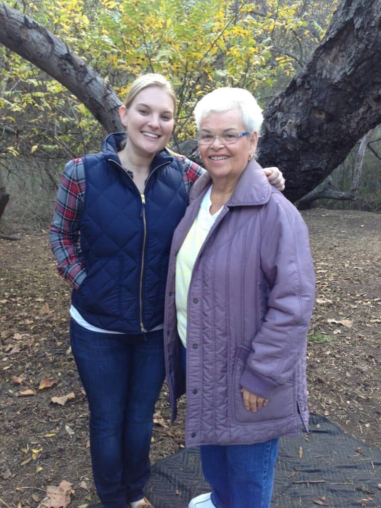 Two white women stand in front of a tree with their arms around each other. The one on the left is in her late 20s and is wearing dark jeans with a plaid shirt and a blue puffer vest. The one on the right is in her 70s and is wearing jeans with a purple puffer coat. They are both grinning.