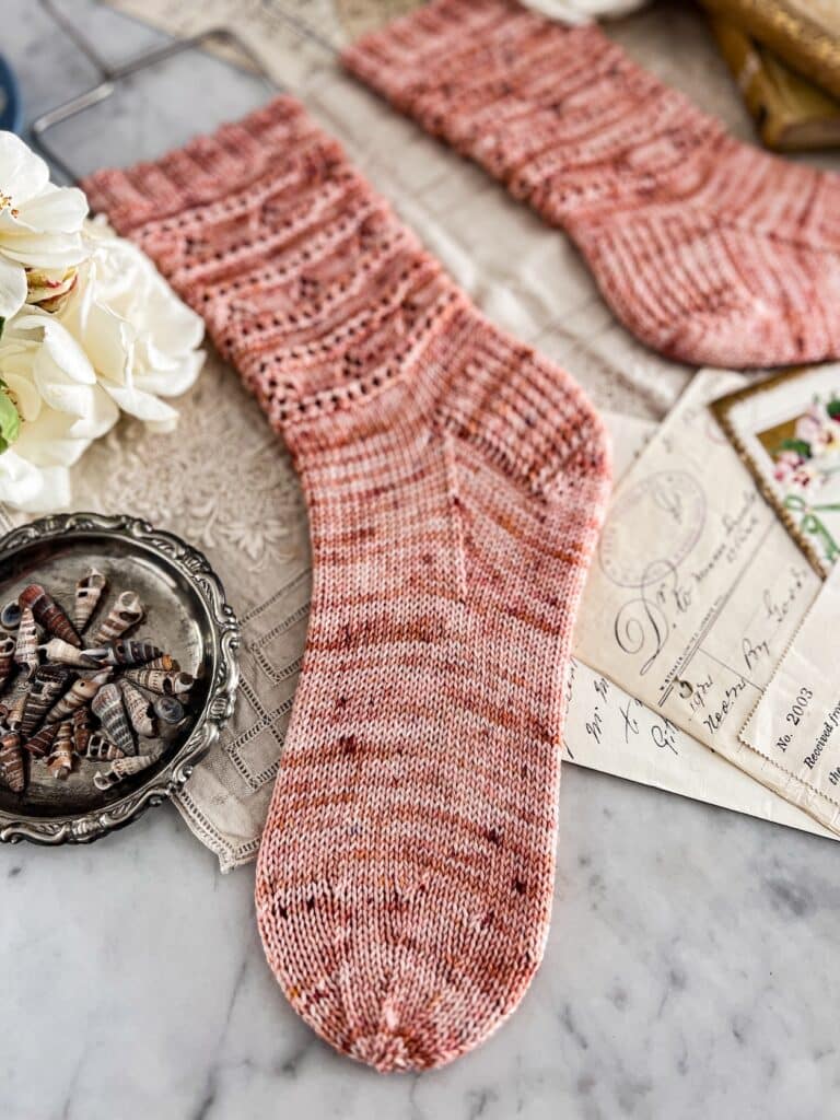 A close-up on the round toe of a pair of pink handknit socks. In the background are some white roses and antique paper ephemera.