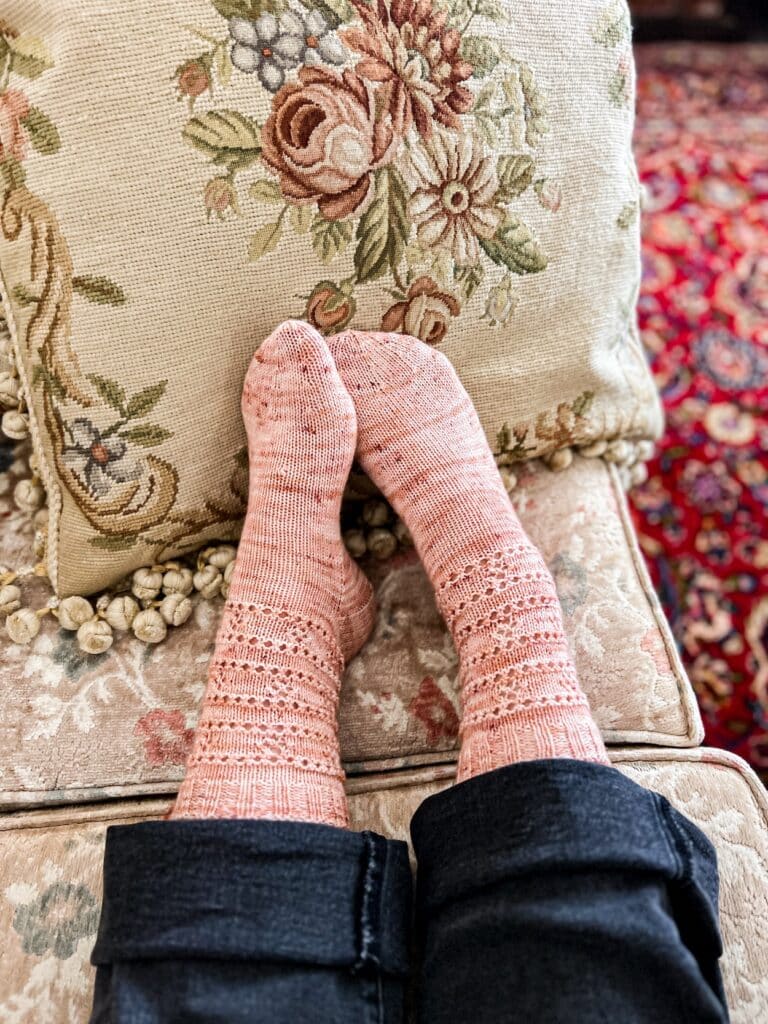 A pair of feet wearing pink handknit socks rest on a needlepoint cushion. The feet are slightly crossed at the toes. The socks have simple eyelet patterns on the leg and a round toe.