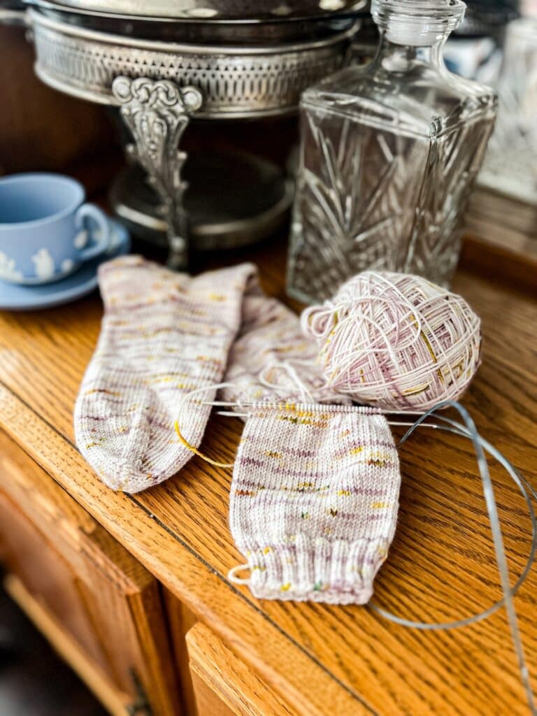 A pair of handknit socks in progress are laid out on a golden oak china hutch. One sock is finished, while the other is still on the needles. They are knit in plain stockinette stitch using a lavender and yellow speckled yarn. Behind them are some vintage dishes including a cut glass decanter, a silver plated serving dish, and a light blue Jasperware teacup and saucer,