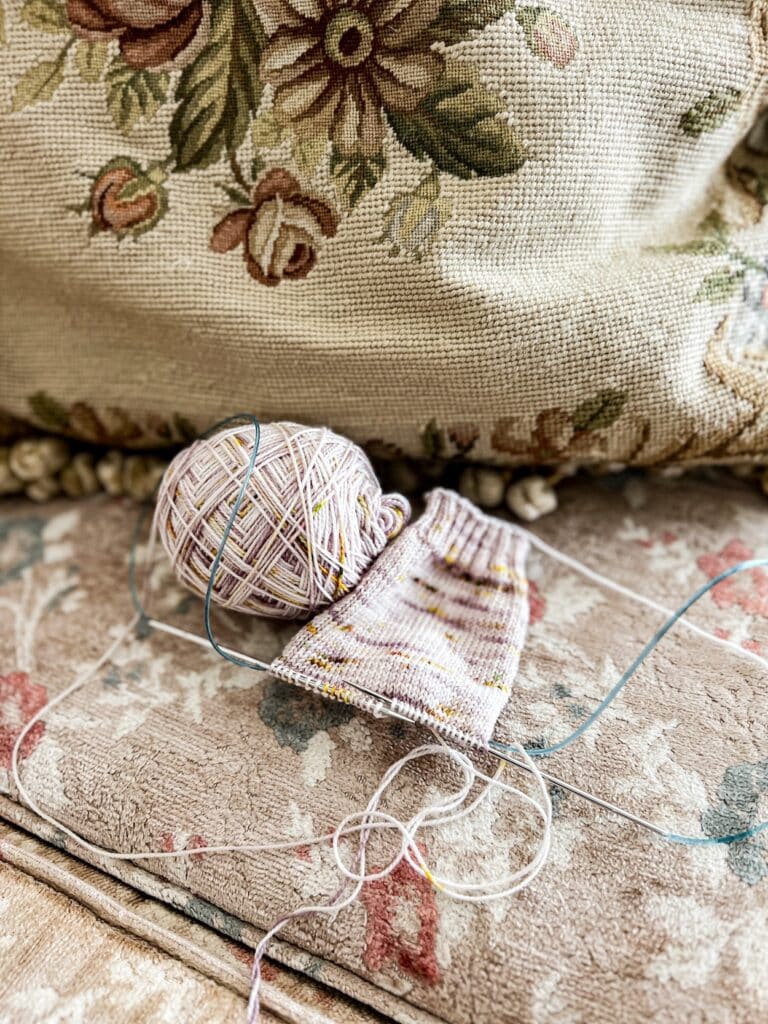 A handknit sock in progress sits on a vintage sofa with worn tan upholstery. The sock is knit in plain stockinette stitch using lavender and yellow speckled yarn. The twisting light blue cords show that the sock is being knit with the magic loop technique.