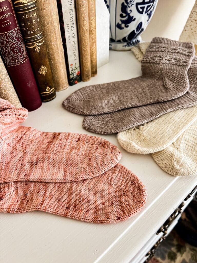 Three pairs of handknit socks--one pink with a round toe, one white with a wedge toe, and one gray with a star toe--are laid out on a white dresser top with some antique books in the background.