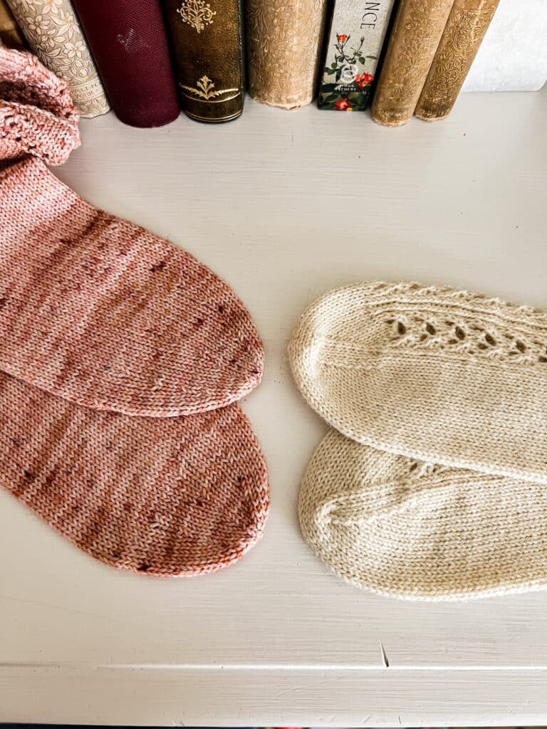 Two pairs of handknit socks, one pink and one white, are laid out on a white dresser with some antique books in the background. The pink socks, on the left, have round toes. The white socks, on the right, have wedge toes. All socks are displayed on their side to show that the wedge toe has very distinct, visible decrease lines, while the round toe does not.