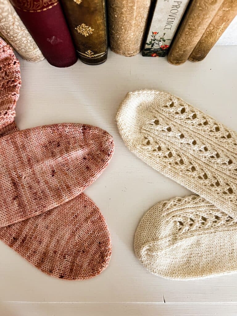 Two pairs of handknit socks, one pink and one white, are laid out on a white dresser with some antique books in the background. The pink socks, on the left, have round toes. The white socks, on the right, have wedge toes. Each pair has one sock displayed on its side and one displayed top-down to show that while the round toe has radial symmetry, the wedge toe does not. This angle also more clearly shows the wedge shape of the wedge toe.