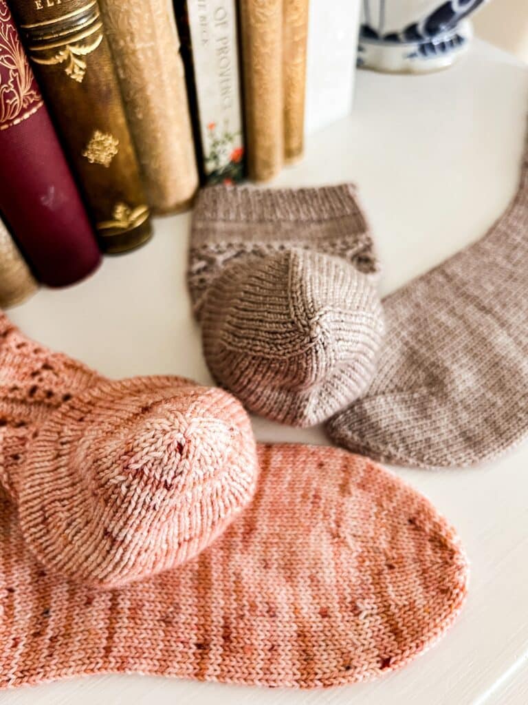 Two pairs of handknit socks, one pink and one gray, are laid out on a white dresser with some antique books in the background. The pink socks, on the left, have round toes. The gray socks, on the right, have star toes. Each pair has one sock displayed on its side and one with the toe shown stuffed and pointing upward as if it were on a foot, to show that the star toe has more distinct and visible decrease lines compared to the round toe when worn.