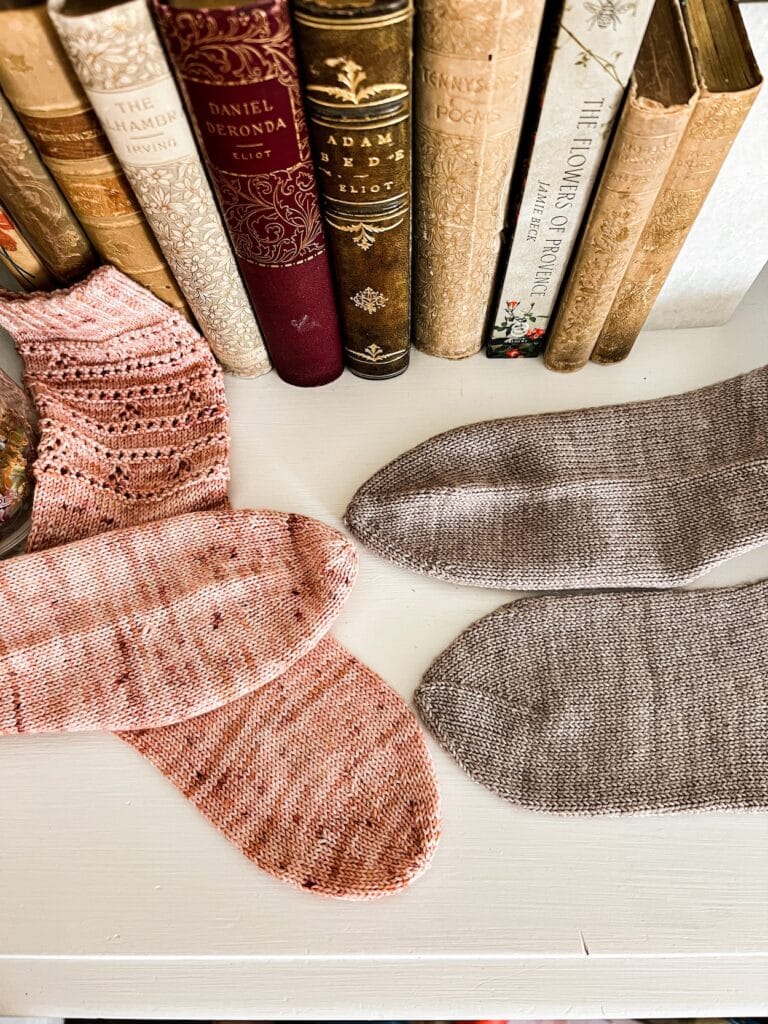 Two pairs of handknit socks, one pink and one gray, are laid out on a white dresser with some antique books in the background. The pink socks, on the left, have round toes. The gray socks, on the right, have star toes. Each pair has one sock displayed on its side and one displayed top-down to show the radial symmetry of both toes.