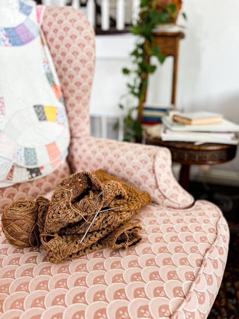 A brown, lacy, handknit sweater in progress rests on a pink wingback chair with a vintage quilt draped over the back of it.