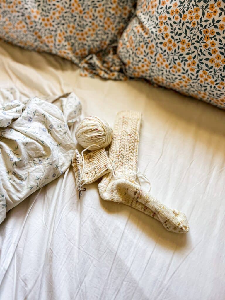 A finished handknit sock and another handknit sock still on the needles rest on an unmade bed surrounded by a floral duvet and floral pillows. The socks are knit in a creamy yarn speckled with faint orange and yellow flecks, and they feature lots of texture using slipped stitches and coin lace.