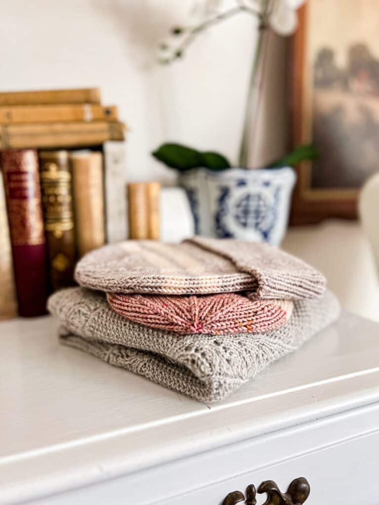 A stack of gray and pink handknits are folded on top of a white dresser with lots of antique books blurred in the background.