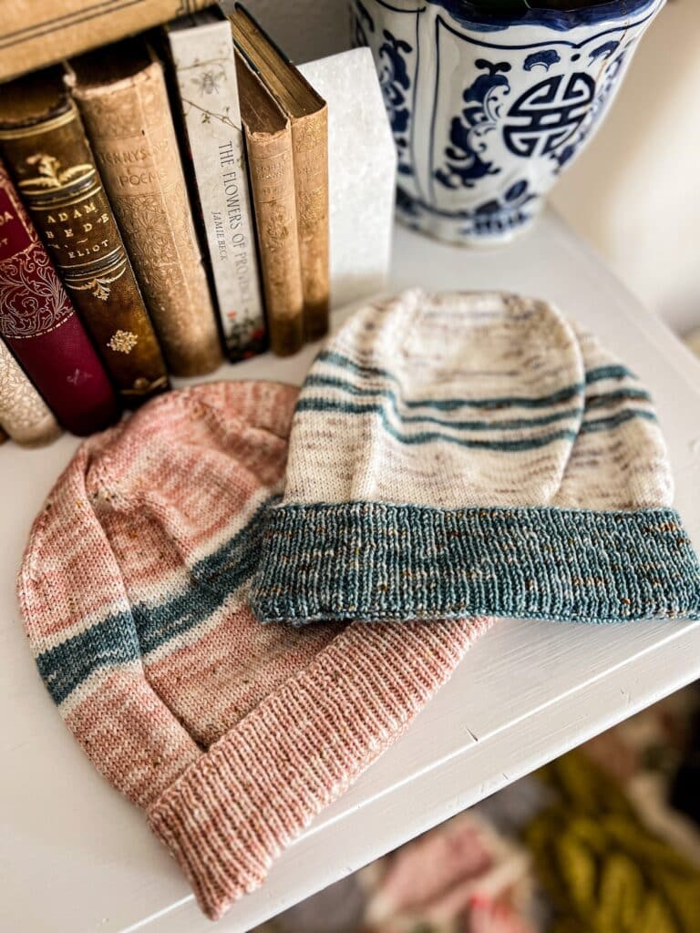 Two stockinette knit hats are laid flat on the top of a white dresser with lots of antique books in the background. The bottom hat is pink with blue and white stripes. The top hat is white with three blue stripes and a blue ribbed brim. Both hats are knit in simple stockinette stitch.
