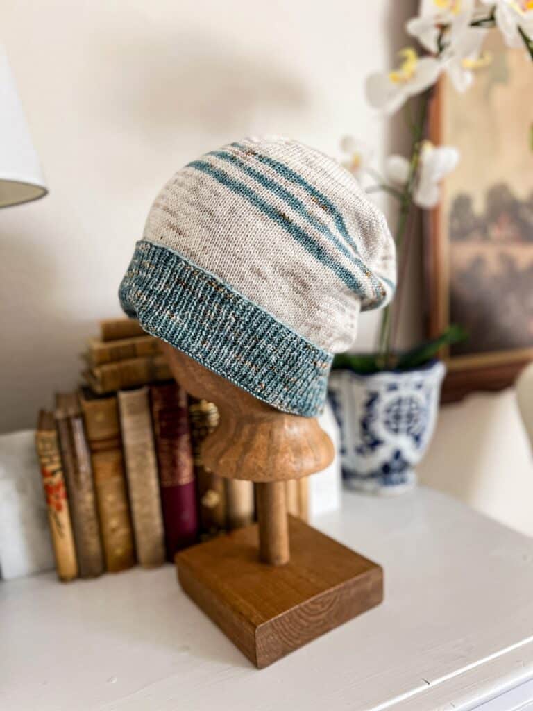 A blue and white striped knit hat with a blue brim is displayed on a brown mannequin head. The hat is slightly slouchy and photographed from the side.