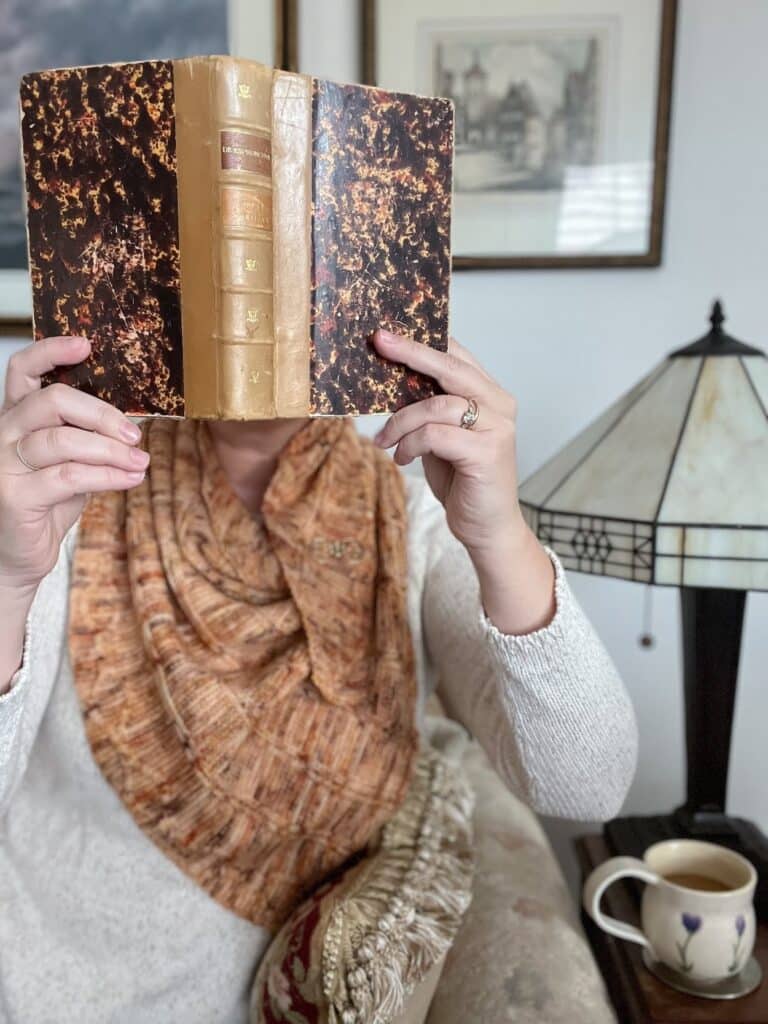 A white woman holds an antique book with a brown leather spine and marbled cover paper. The book is open and covers her face so that only her hands and torso are visible. She's wearing an orange triangle knit shawl and a cream sweater.