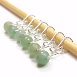 Several green beaded stitch markers with silver clasps dangle from a wooden knitting needle