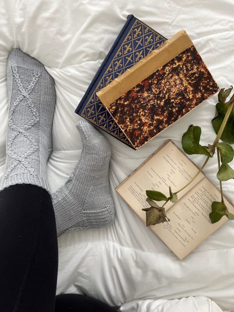A woman's feet wearing a pair of light blue handknit socks rest on top of a white comforter. Next to them are a couple antique books, one of which is open and has some faded hellebores resting on it.