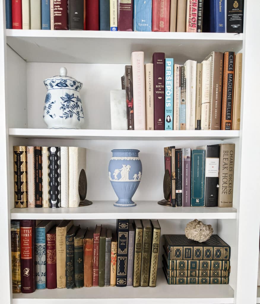 Three white shelves full of books, some vintage and antique, some new. There's also a blue and white Wedgewood Jasperware vase, a hunk of coral, and a blue onion-printed cookie jar.