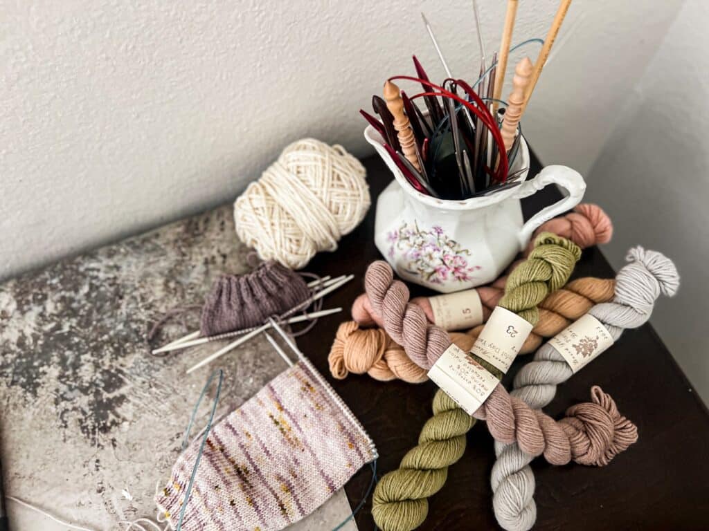 A top-down photo where a white stoneware pitcher full of metal and wooden knitting needles sits on a desk surrounded by pastel mini skeins of yarn and a couple knitting projects in progress on metal and plastic needles.