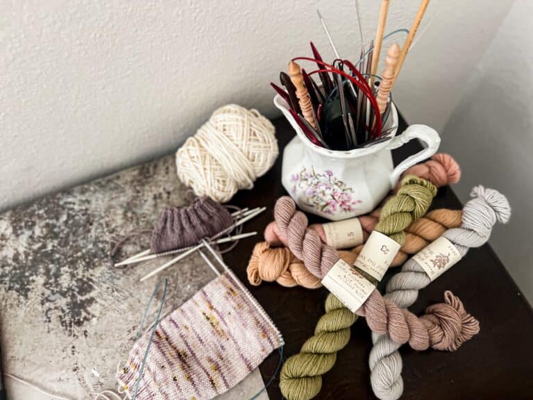 A top-down photo where a white stoneware pitcher full of metal and wooden knitting needles sits on a desk surrounded by pastel mini skeins of yarn and a couple knitting projects in progress on metal and plastic needles.