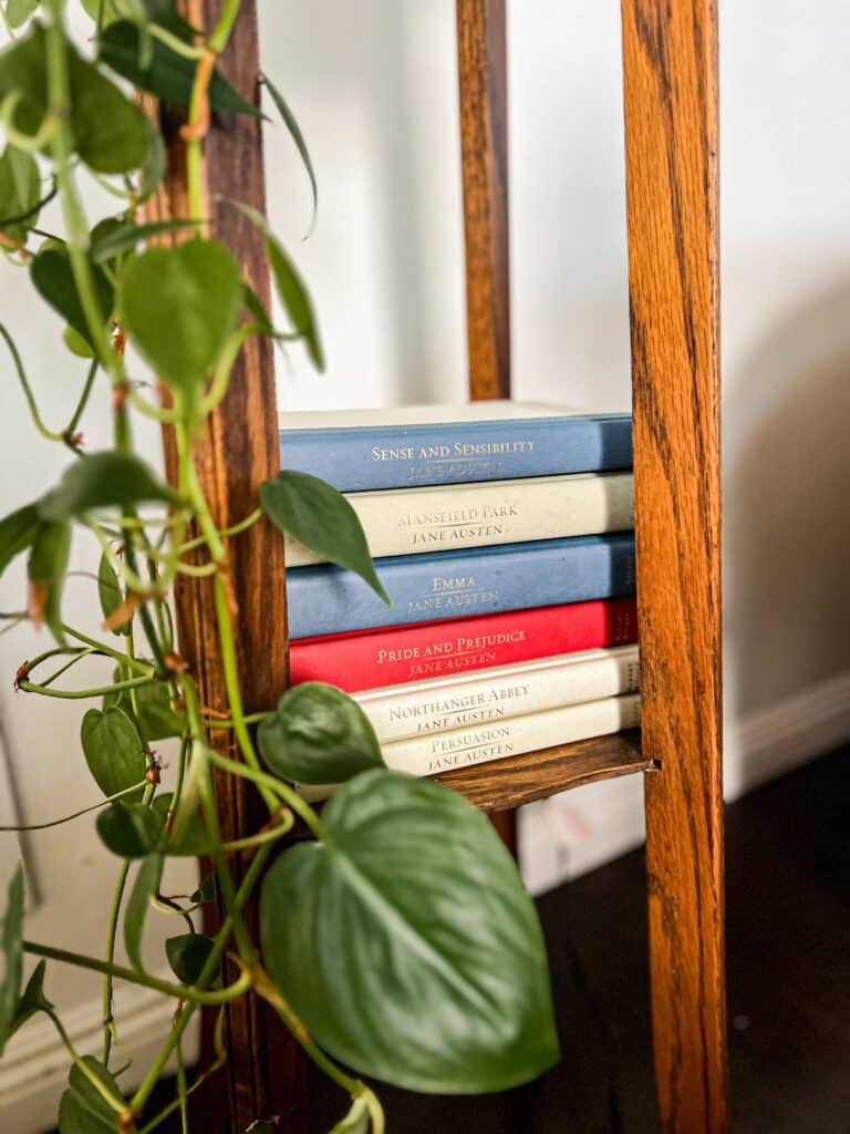 A stack of Jane Austen books turned on their side fills the bottom level of a plant stand. On the left and in the foreground are lots of glossy green leaves from a pothos plant.