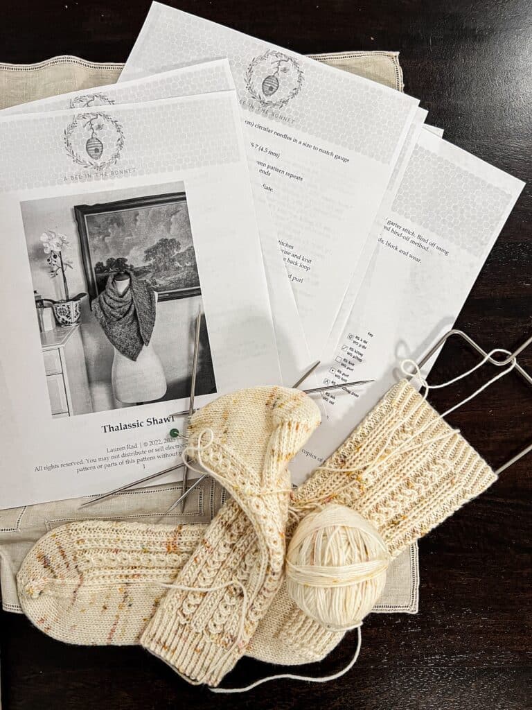 A top-down photo showing the many pages of a relatively simple knitting pattern and a pair of handknit cream-colored socks. One sock is still in progress on a pair of double-pointed needles.