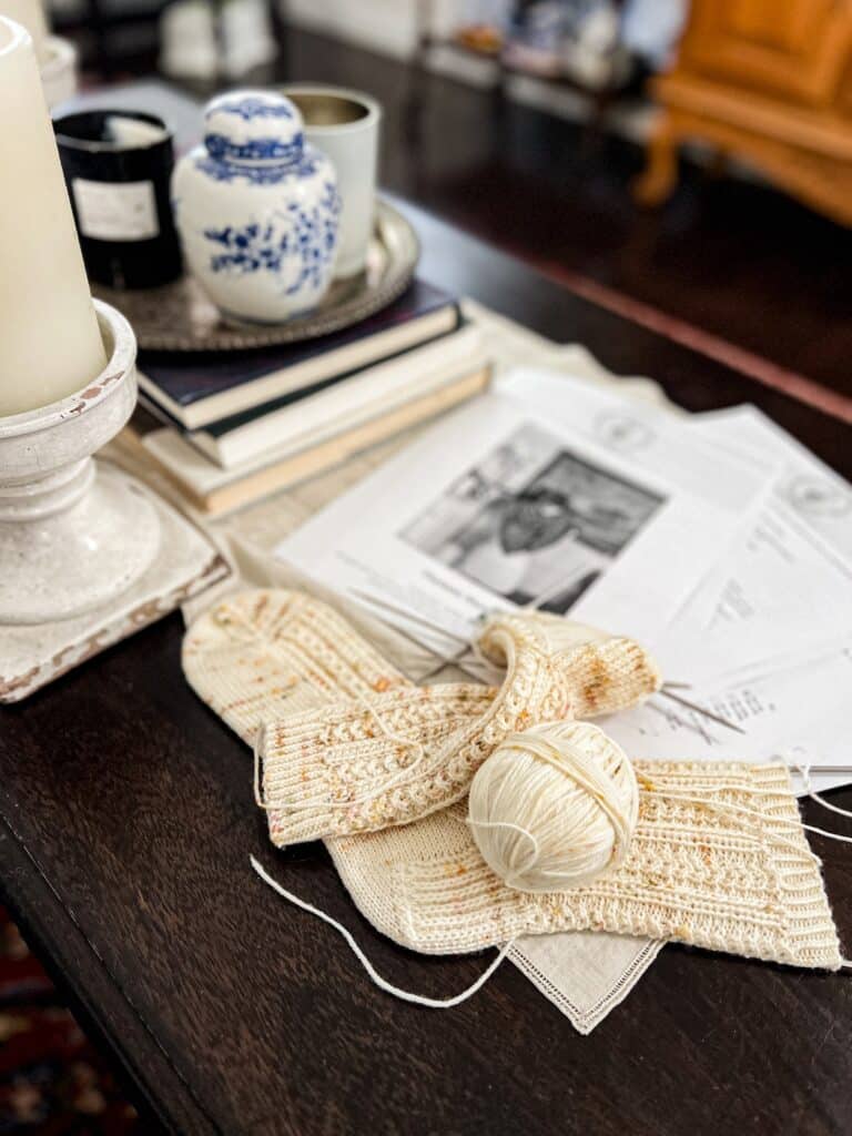 A photo taken from a low angle showing a cream-colored knit sock in progress and a ball of yarn. Blurred in the background are many pages of a knitting pattern and some vintage tabletop decor (candles, a silver tray, books, etc.).