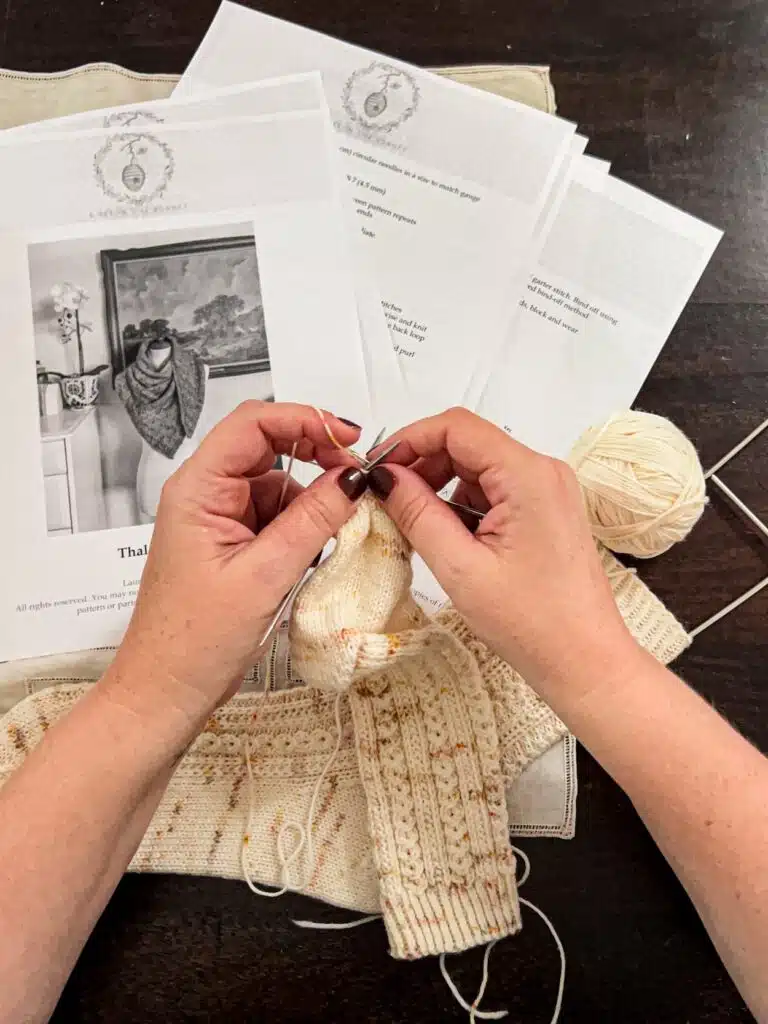 A top-down photo showing a white woman's hands knitting a partially finished sock in cream yarn. Spread on the table in the background are the many pages of a relatively simple knitting pattern.
