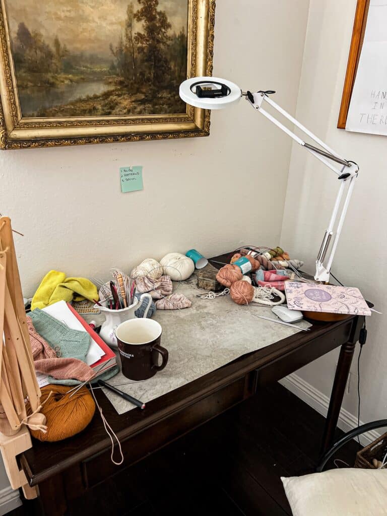 A dark wooden desk with long, spindly legs is almost completely covered in half-finished knitting projects, notebooks, hanks of yarn, knitting needles, and lots of other accessories related to designing knitting patterns.