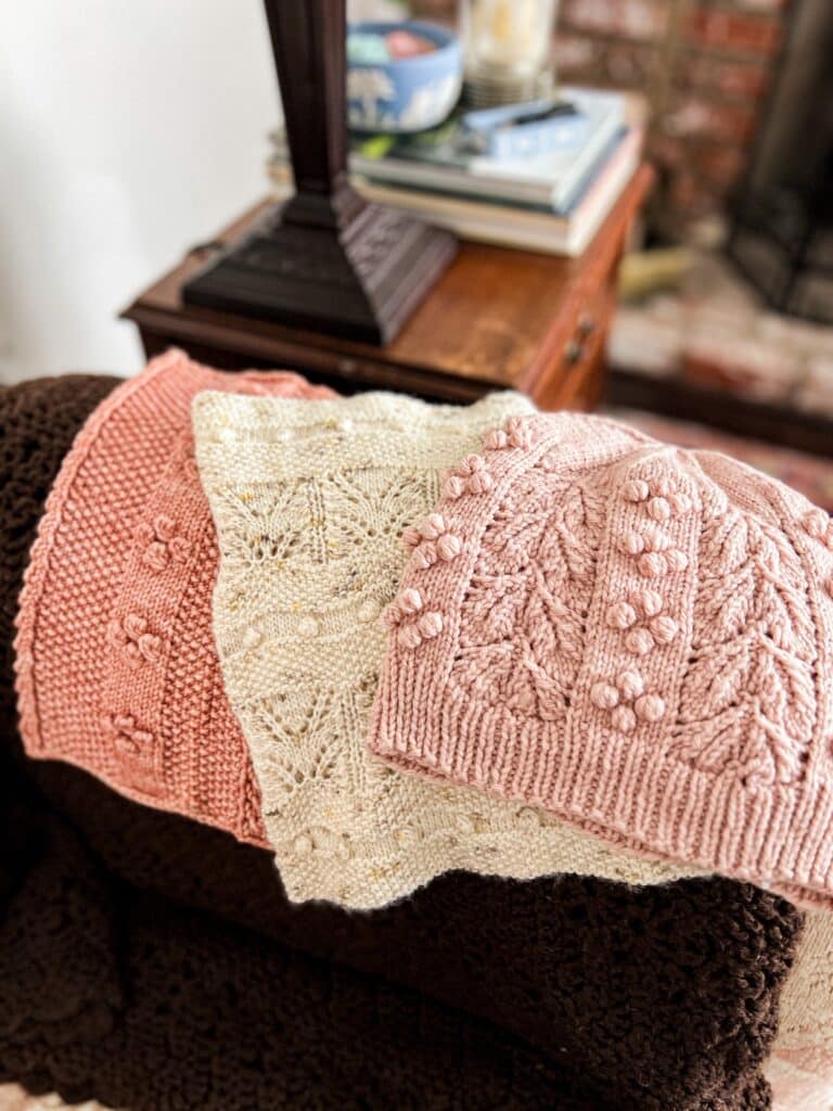 Three handknit accessories (a pink cowl, a cream cowl, and a pink hat) are all laid out on top of a dark brown crocheted blanket. Each of the knit accessories features bobbles as a decorative element.
