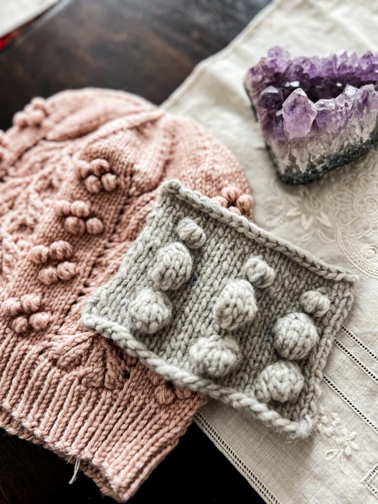 A gray swatch of knit fabric showing three different bobble styles sits in the center foreground of the photo atop a pink knit hat that also has bobbles on it.