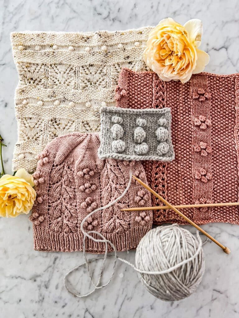 A top-down photo of a pile of pink and cream knit accessories with a gray swatch in the middle. All of the knit items have bobbles on them, including the swatch. They're surrounded by gray yarn, wooden knitting needles, and yellow roses.