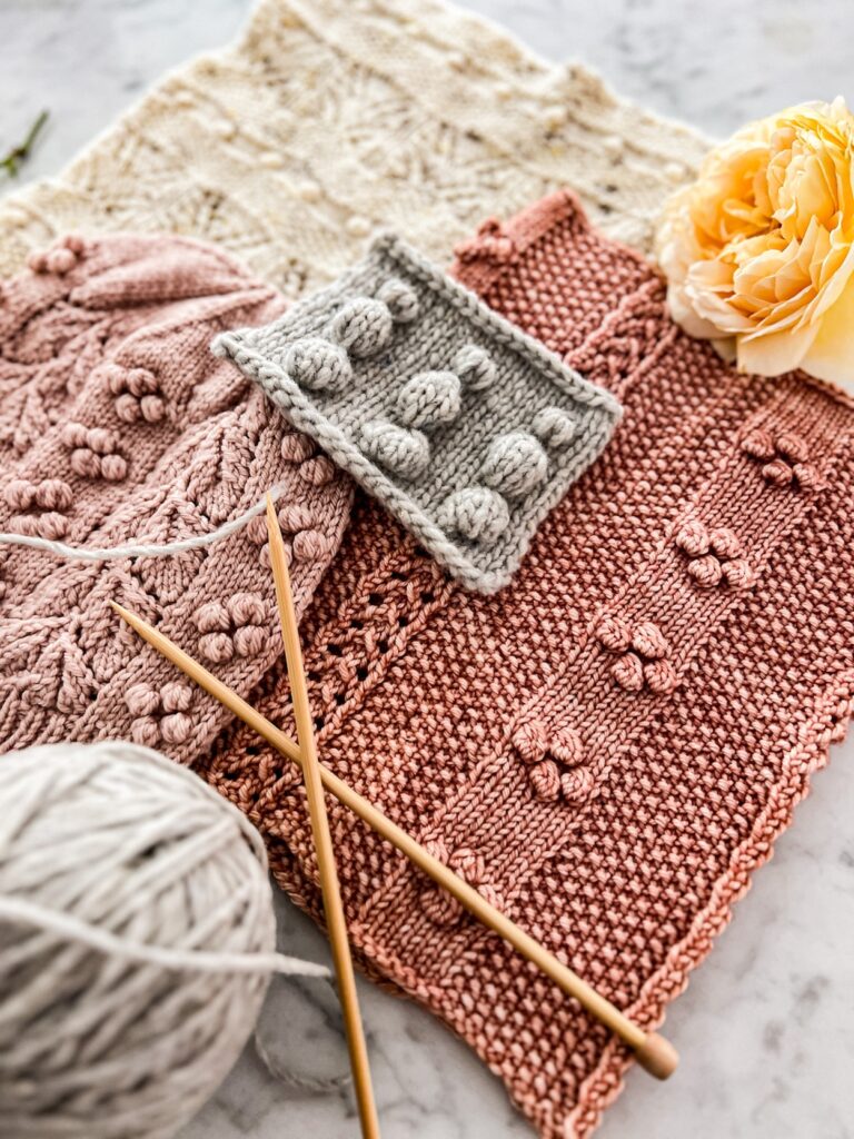 A pile of pink and cream knit accessories sit on a marble countertop surrounded by yellow roses, gray yarn, and wooden knitting needles. In the center of the pile is a gray knit swatch showing three different types of knit bobbles.