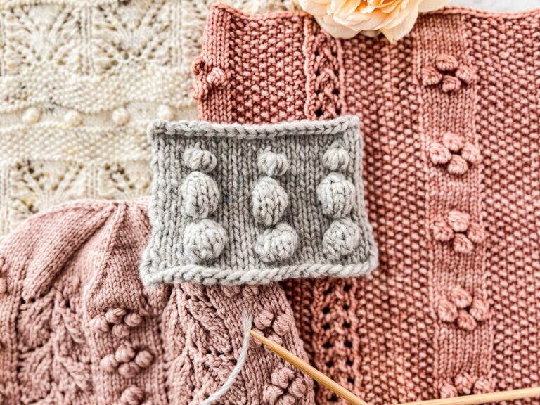 A top-down photo of a pile of pink and cream knit accessories with a gray swatch in the middle. All of the knit items have bobbles on them, including the swatch.
