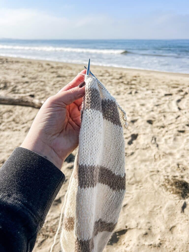 A white woman's left hand holds up a white and tan knit scarf in progress. In the background are lots of sand and some soft waves at the beach.