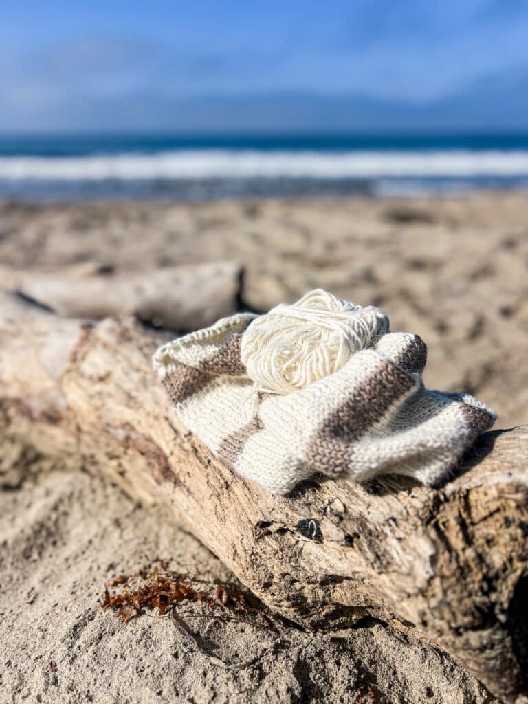 A half-finished knit scarf with tan and white stripes sits on a driftwood log at the beach. Blurred in the background is lots of sand and some soft blue waves.