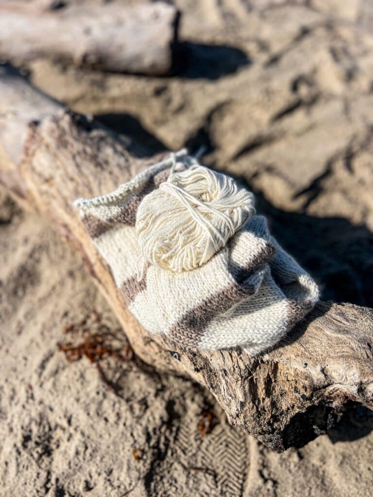 A half-finished knit scarf with tan and white stripes sits on a driftwood log at the beach, surrounded by sand. The photograph is taken from the top down.