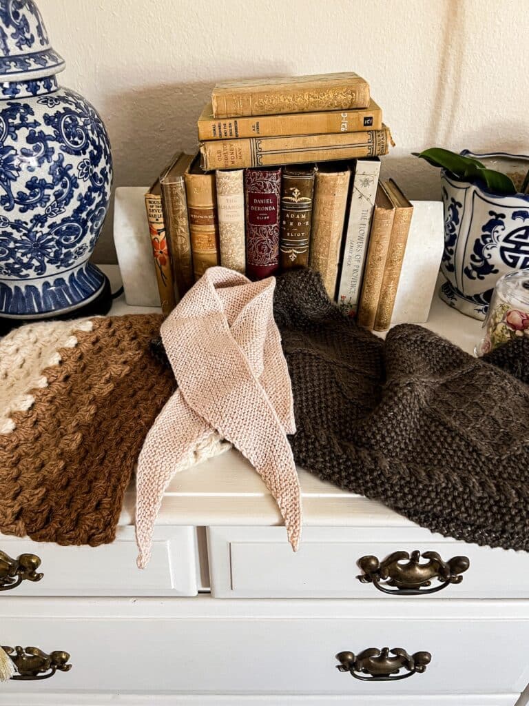 A pile of items made with alpaca sit on top of a white dresser. From left to right are a crocheted cowl in three different natural colors of alpaca, a pink knit scarflet in plain garter stitch, and a brown knit scarf with lots of cables and seed stitch.