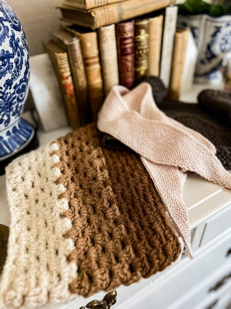 A close-up on a crocheted cowl made with three different natural colors of alpaca. Blurred in the background are a pink scarflet, a brown scarf, and lots of antique books.