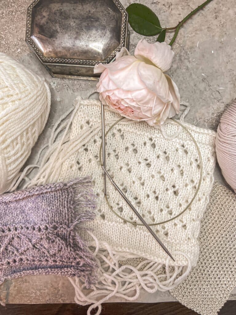 A top-down photo showing two swatches, one with the floats cut and one with the floats intact (the white swatch on the bottom). They're surrounded by more yarn, some metal knitting needles, a silver trinket box, a pink rose, and a seed stitch swatch knit flat.