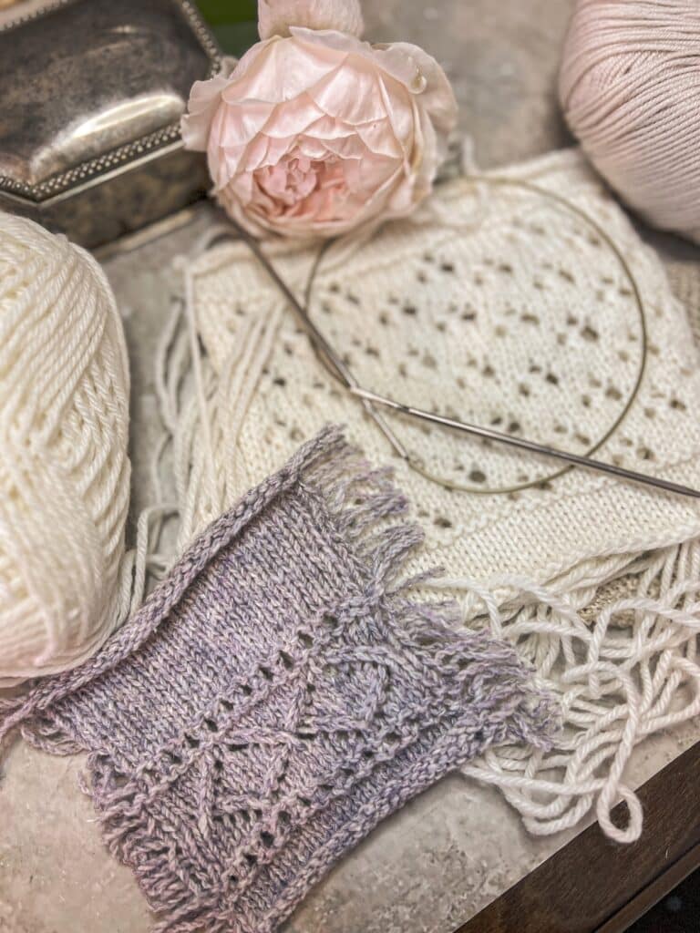 A low-angle photo of a gray speed swatch with its floats cut to a short fringe. Blurred in the background is a white speed swatch with its floats intact, along with a pink rose, a silver trinket box, and some more yarn.