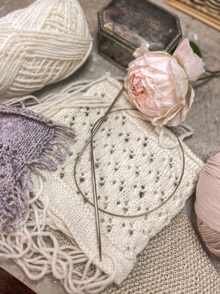A low-angle photo of a white speed swatch with its floats intact. A set of metal knitting needles rests on top of the swatch. A gray swatch with its floats cut is just visible at the left edge of the photo.